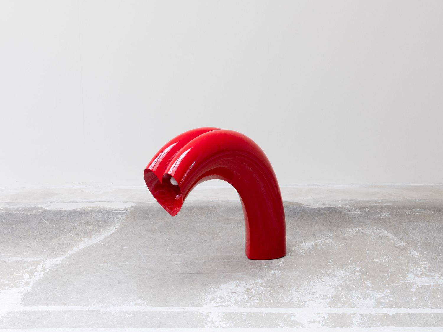 Glossy red arched floor lamp curving over like a candy cane, tubular form dipping to reveal its light at the front tip, on bare concrete — Kimy Gringoire BIGFLOW FLOOR LAMP