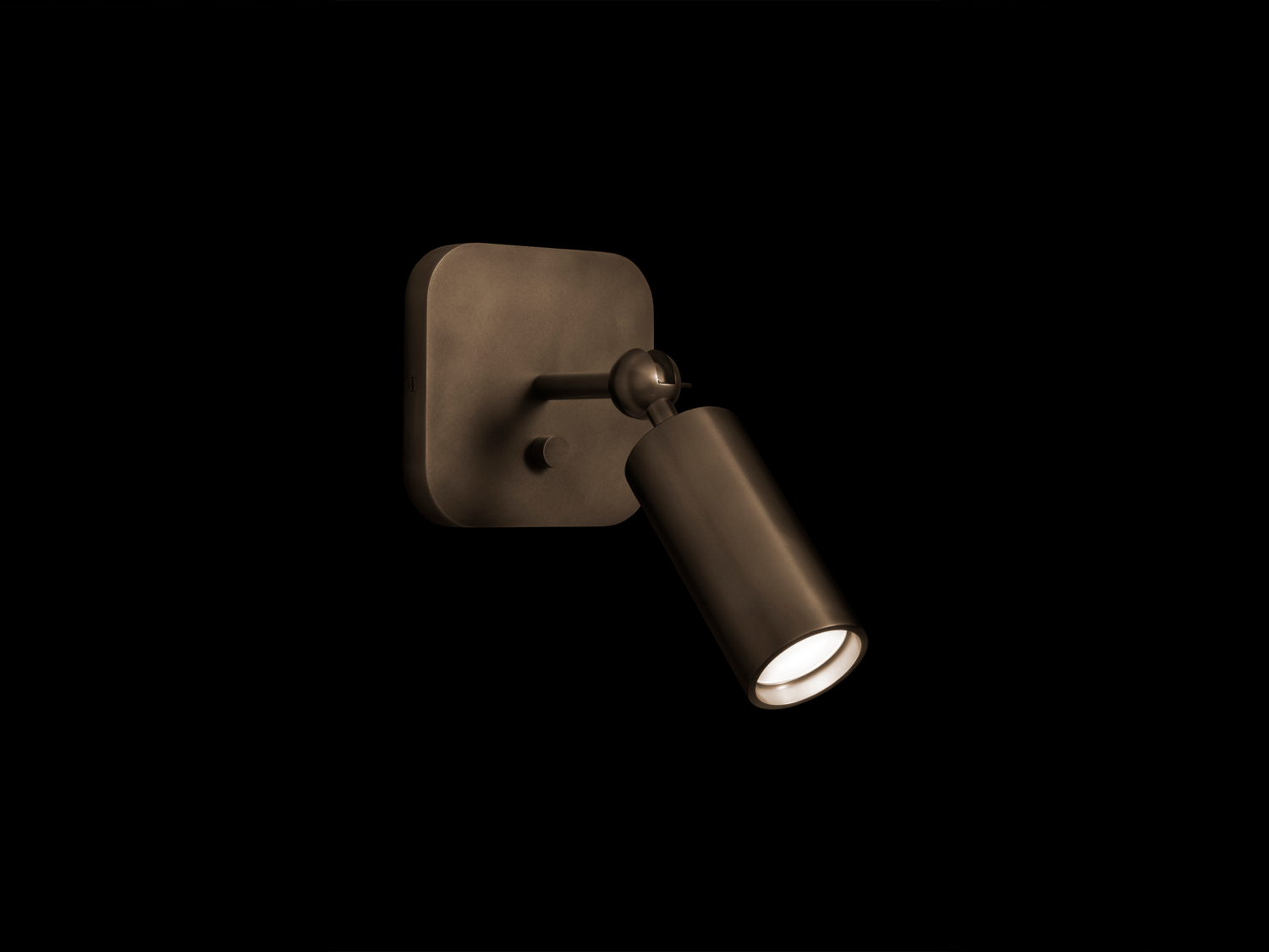 Wall sconce with squared backplate and adjustable cylindrical spotlight shade in dark bronze, glowing lens angled downward on black — Apparatus Studio Cylinder Sconce