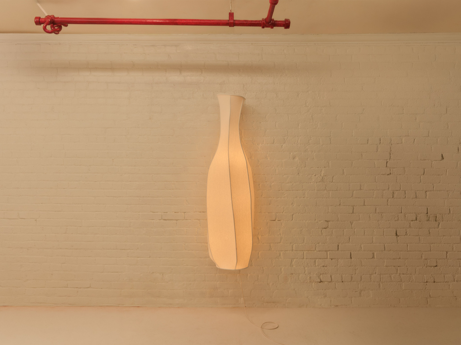 Tall softly glowing bottle-shaped fabric floor lamp against a whitewashed brick wall, red pipe crossing above — Marrow Project Lamp n. 6