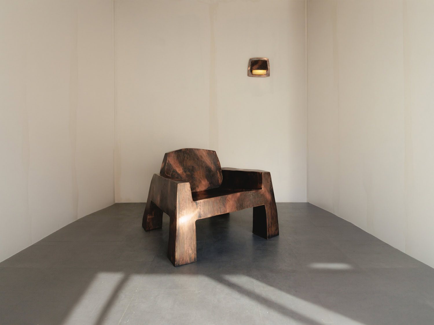Sculptural armchair in dark scorched wood with blocky angular arms and legs, beside a small wall light in a bare plaster room — Nick Valentijn Armchair