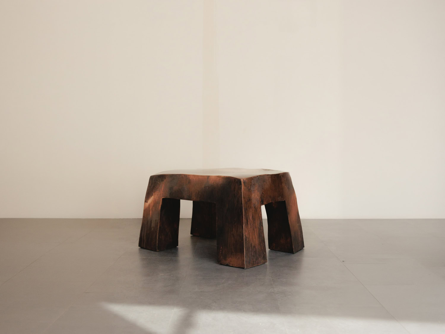 Low sculptural stool in dark scorched wood with a mottled copper-brown finish and four splayed tapering legs, on grey stone floor — Nick Valentijn Ottoman