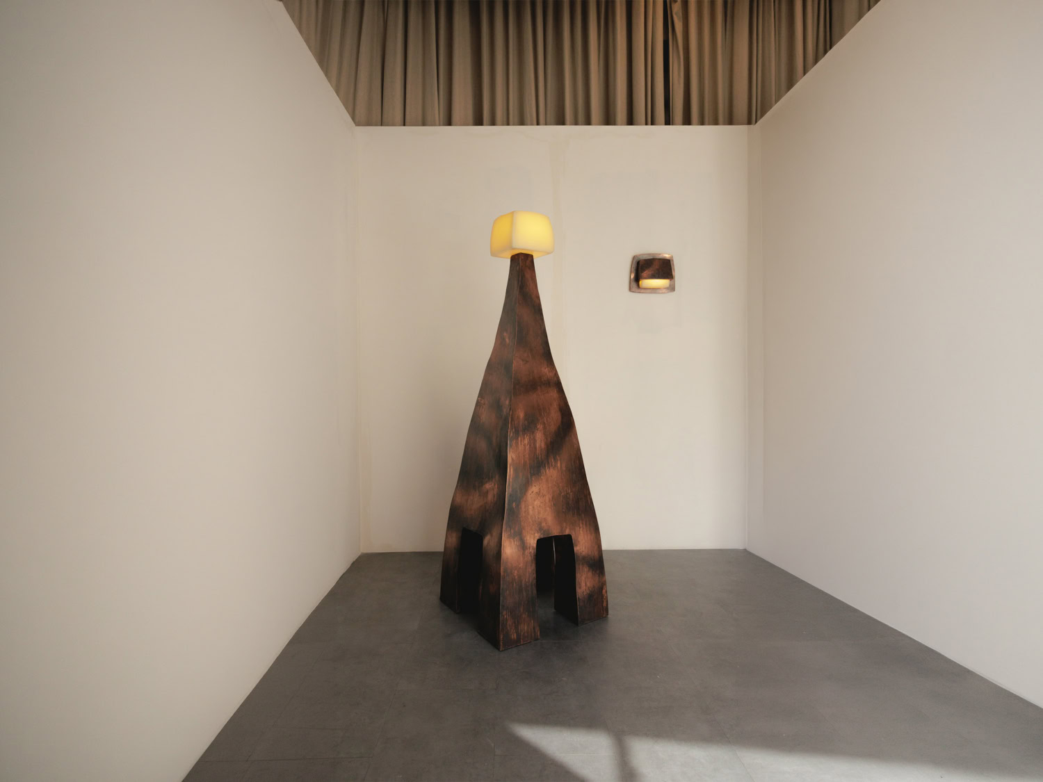 Tall tapering floor lamp on splayed legs in scorched mottled copper, topped by a glowing rounded cube shade — Nick Valentijn Standing Lamp