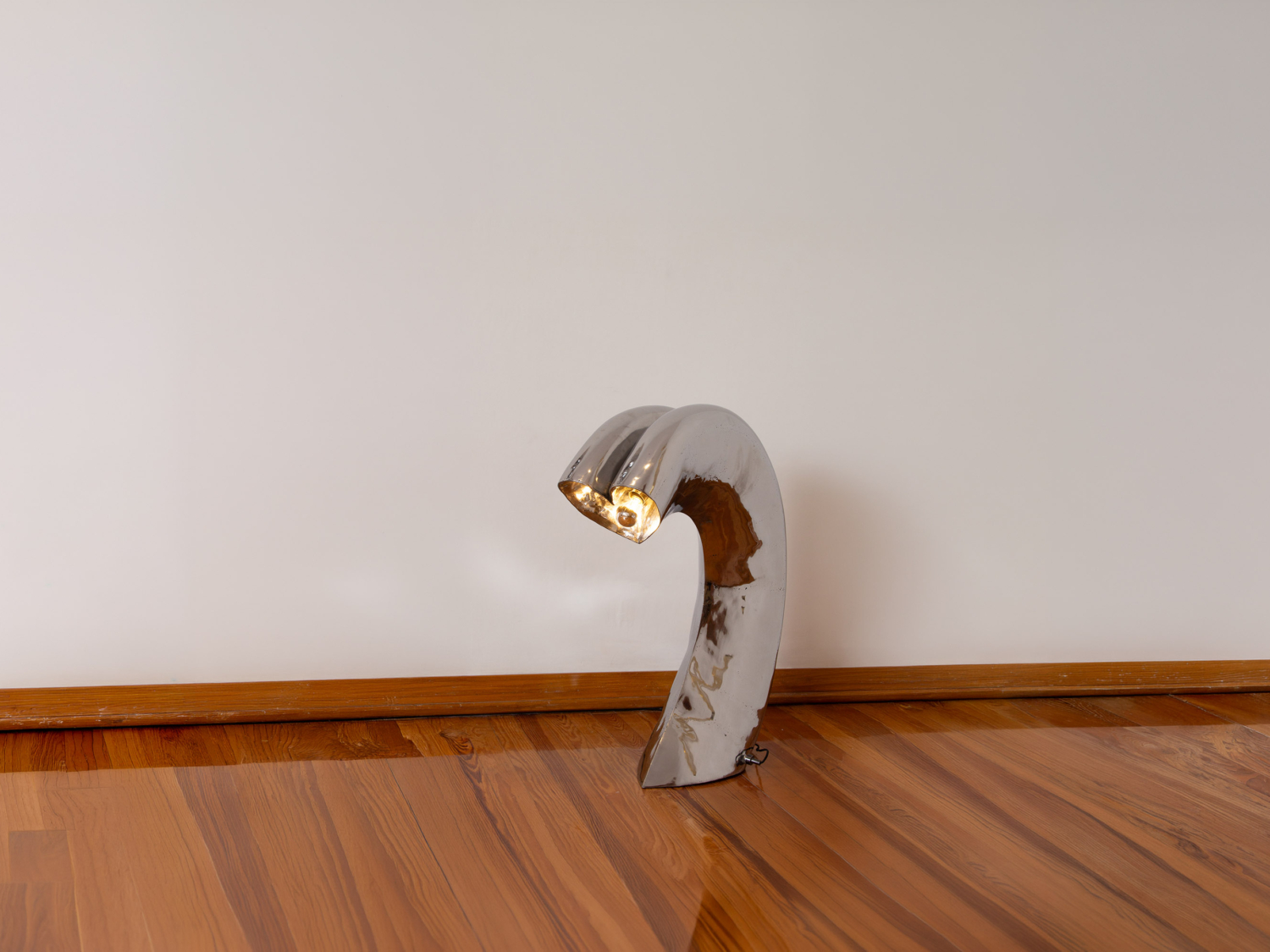 Curved chromed-metal floor lamp arcing over like a question mark, its glowing head resting near a warm wooden floor — Kimy Gringoire BIG FLOW FLOOR LAMP