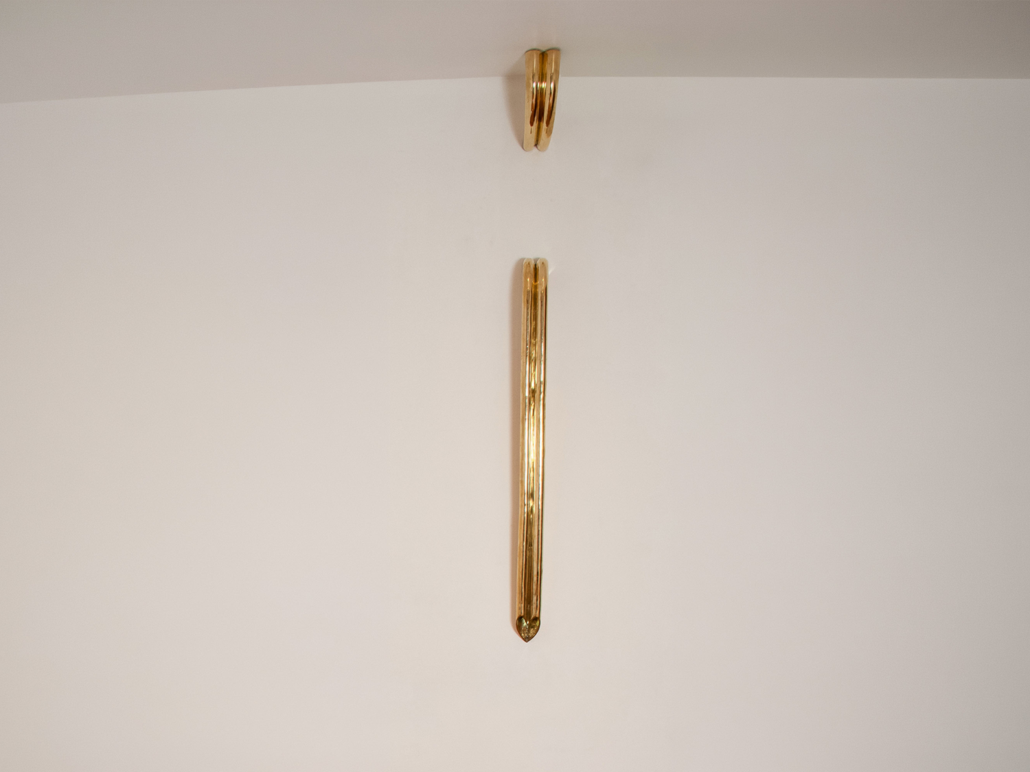 Slim polished brass wall light, a long vertical tube tapering to a point below a small looped ceiling bracket on a pale wall — Kimy Gringoire RIOLOVE GRANDE PENDING