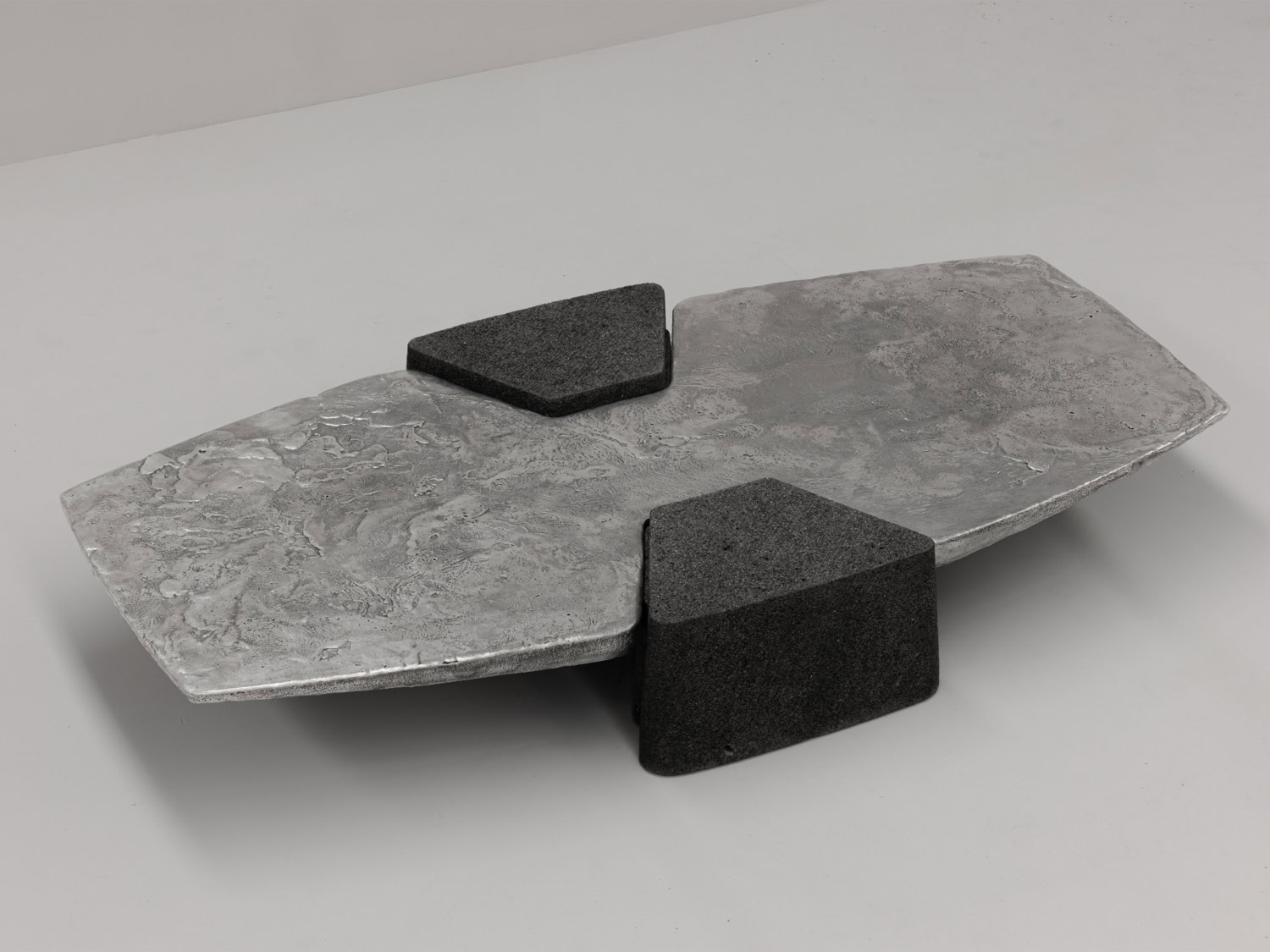 Low coffee table pairing a textured cast-aluminium slab top with two wedge-shaped charcoal volcanic stone bases — EWE Studio EXHUMA Coffee Table