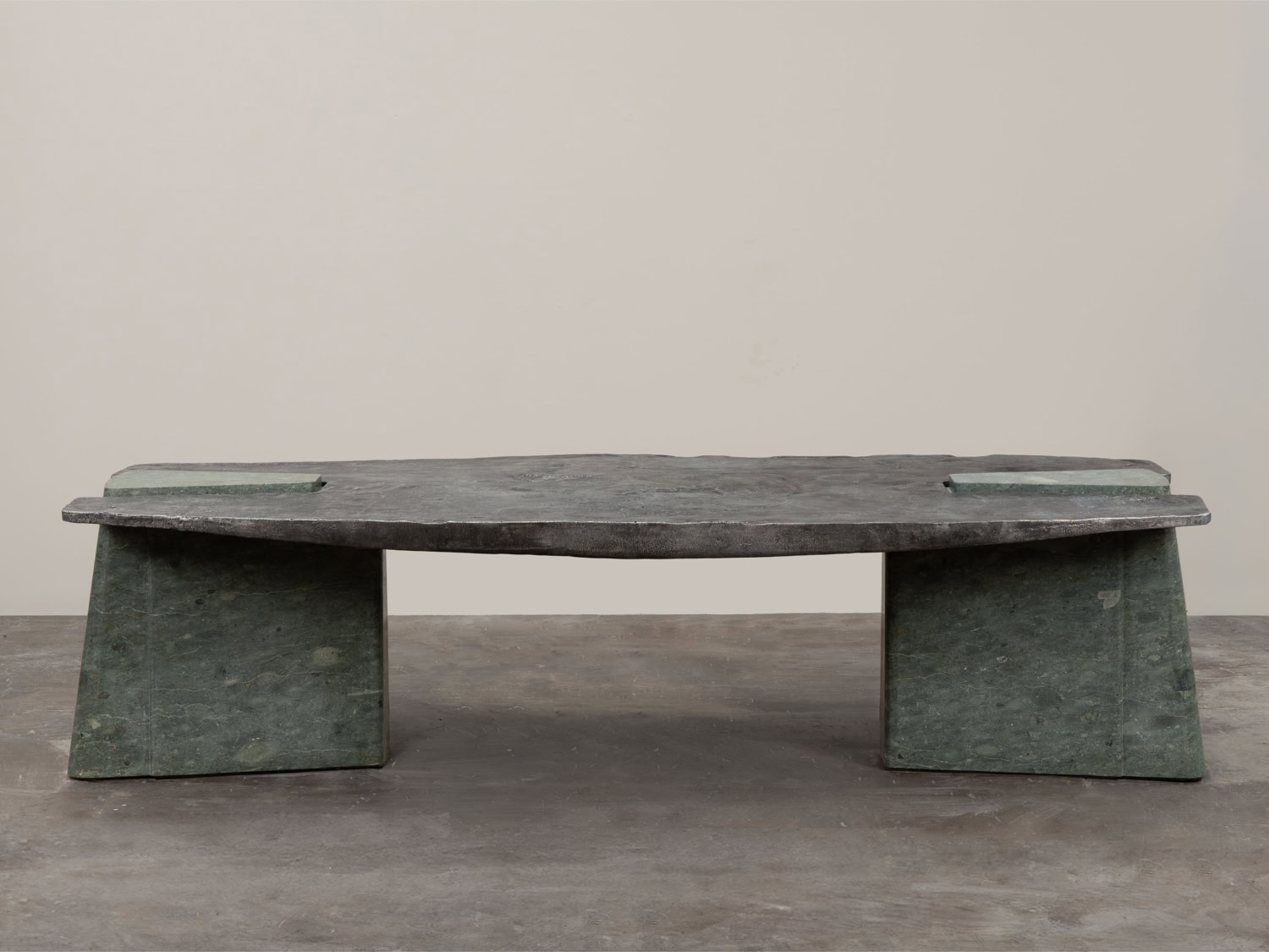 Low dining table with a dark cast-metal top and two trapezoidal green stone block legs, on a concrete floor — EWE Studio Exhuma Dining Table