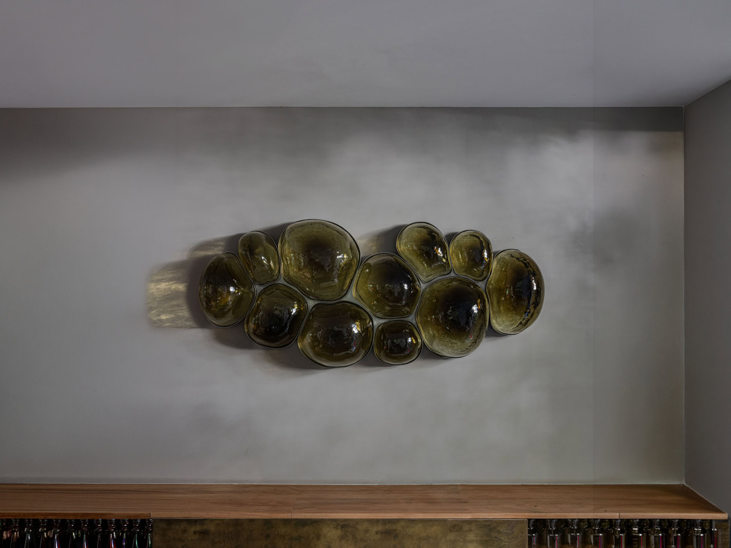 Cluster of glossy olive-green blown-glass bubbles forming a wall-mounted relief above a timber bar, each orb catching light — EWE Studio MAGMA LARGE GREEN